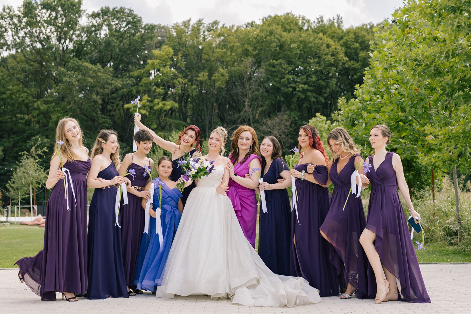 Large bridal party poses in coordinated purple and navy dresses in outdoor garden setting.