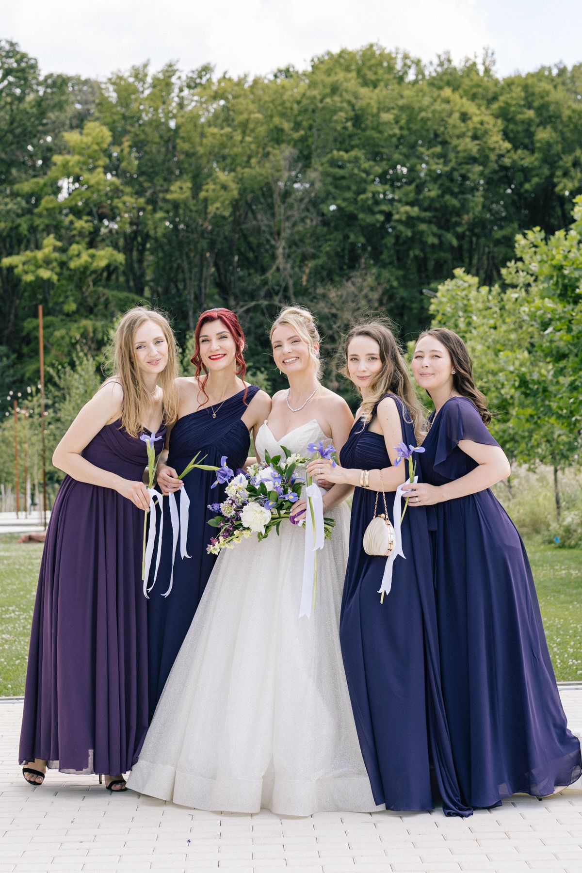 Bridal party in navy and purple dresses poses together in outdoor garden setting.