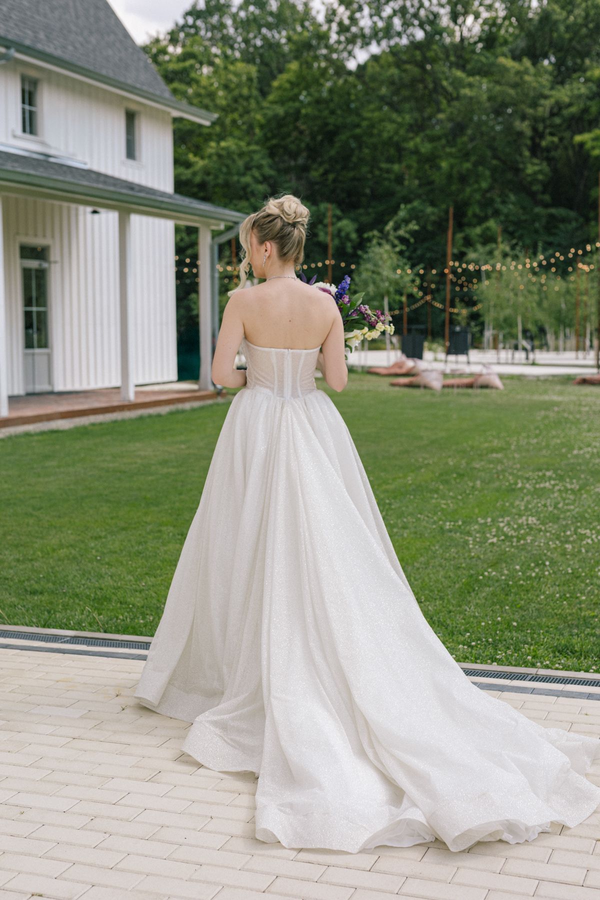 A white wedding dress with a long train photographed from behind on a manicured lawn with string lights.