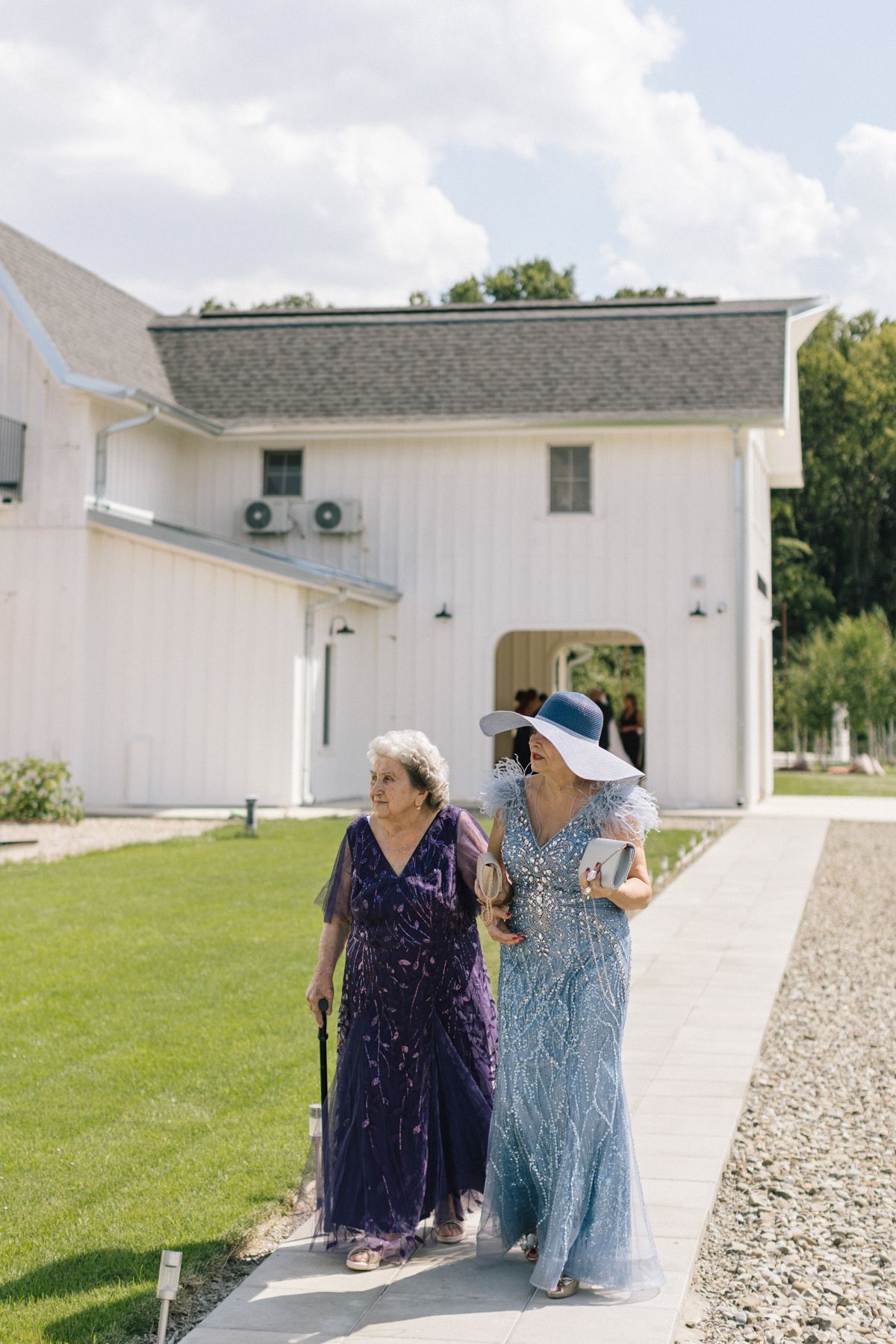 Two guests in formal dresses walking along a path outside a white building.