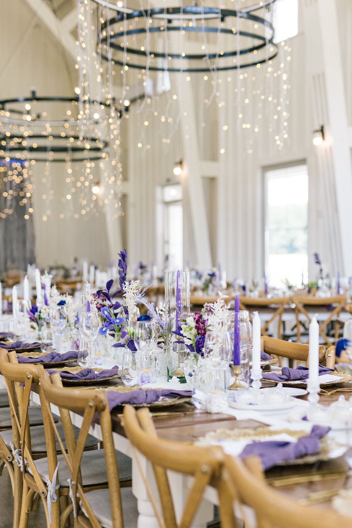Wedding reception table decorated with purple flowers and candles under twinkling lights.
