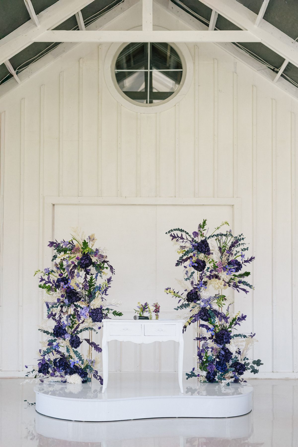 A white wedding ceremony backdrop with two large purple and white floral arrangements.