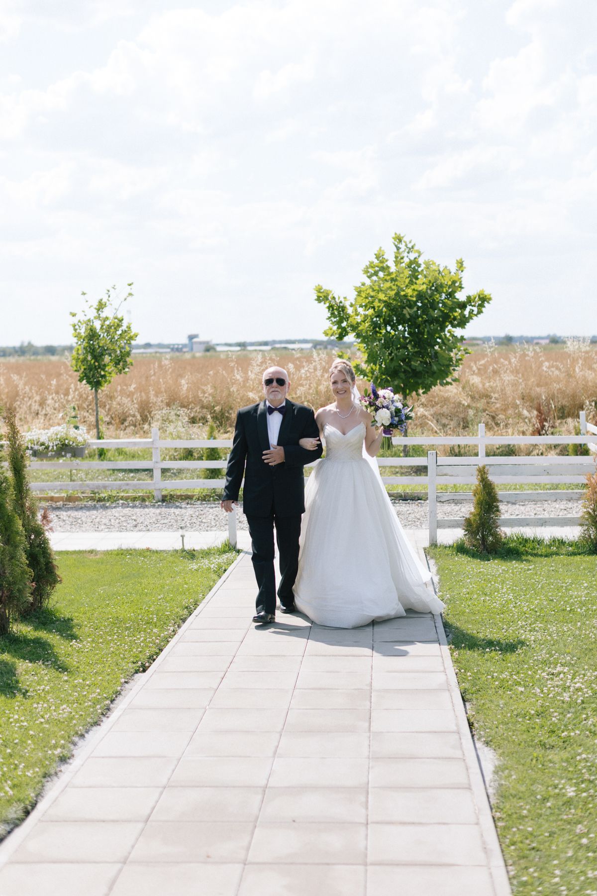 A wedding couple walks down a garden pathway lined with trees and greenery on a sunny day.