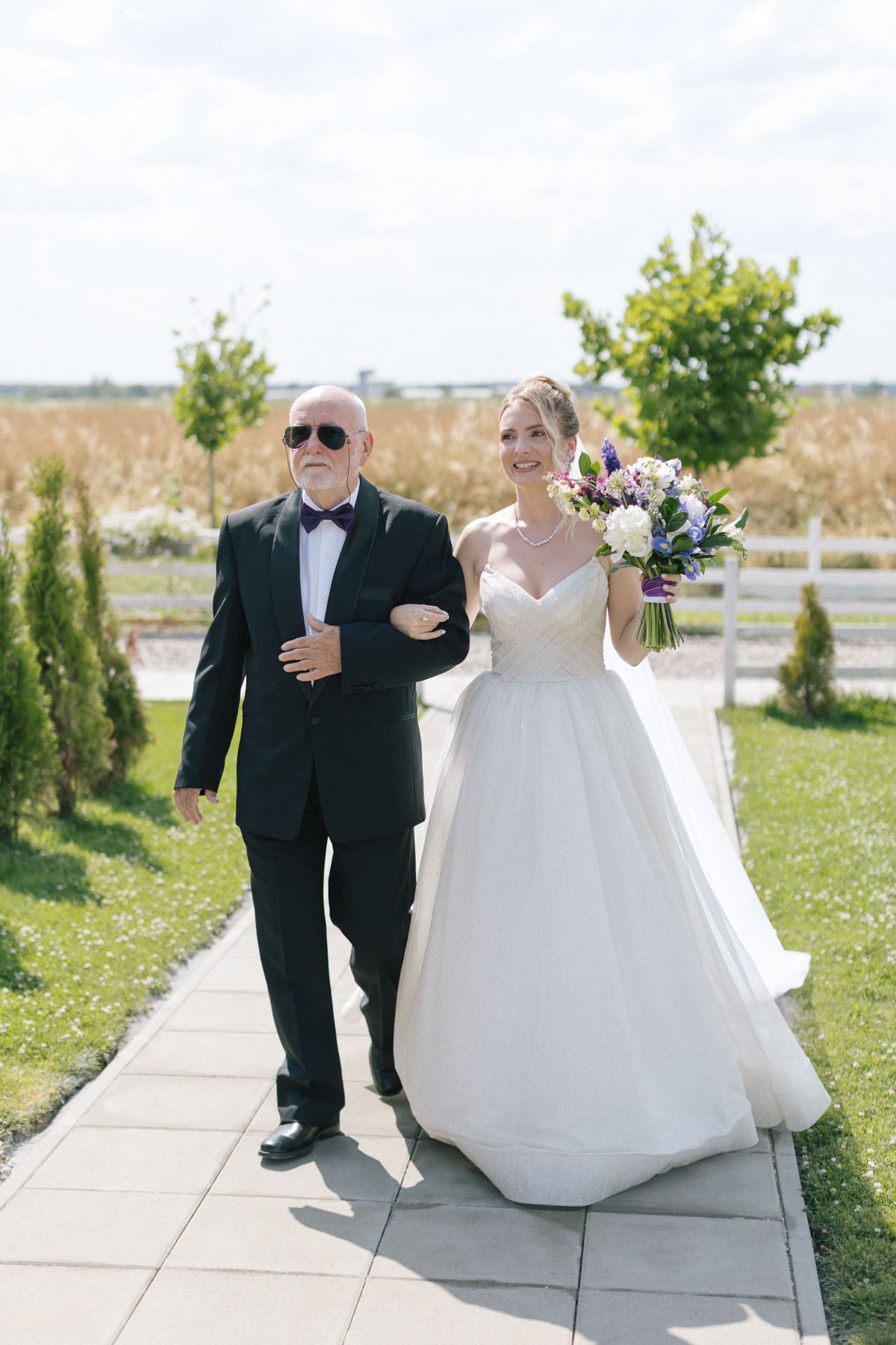 A bride in a white gown walks down an outdoor path with an escort in formal attire.