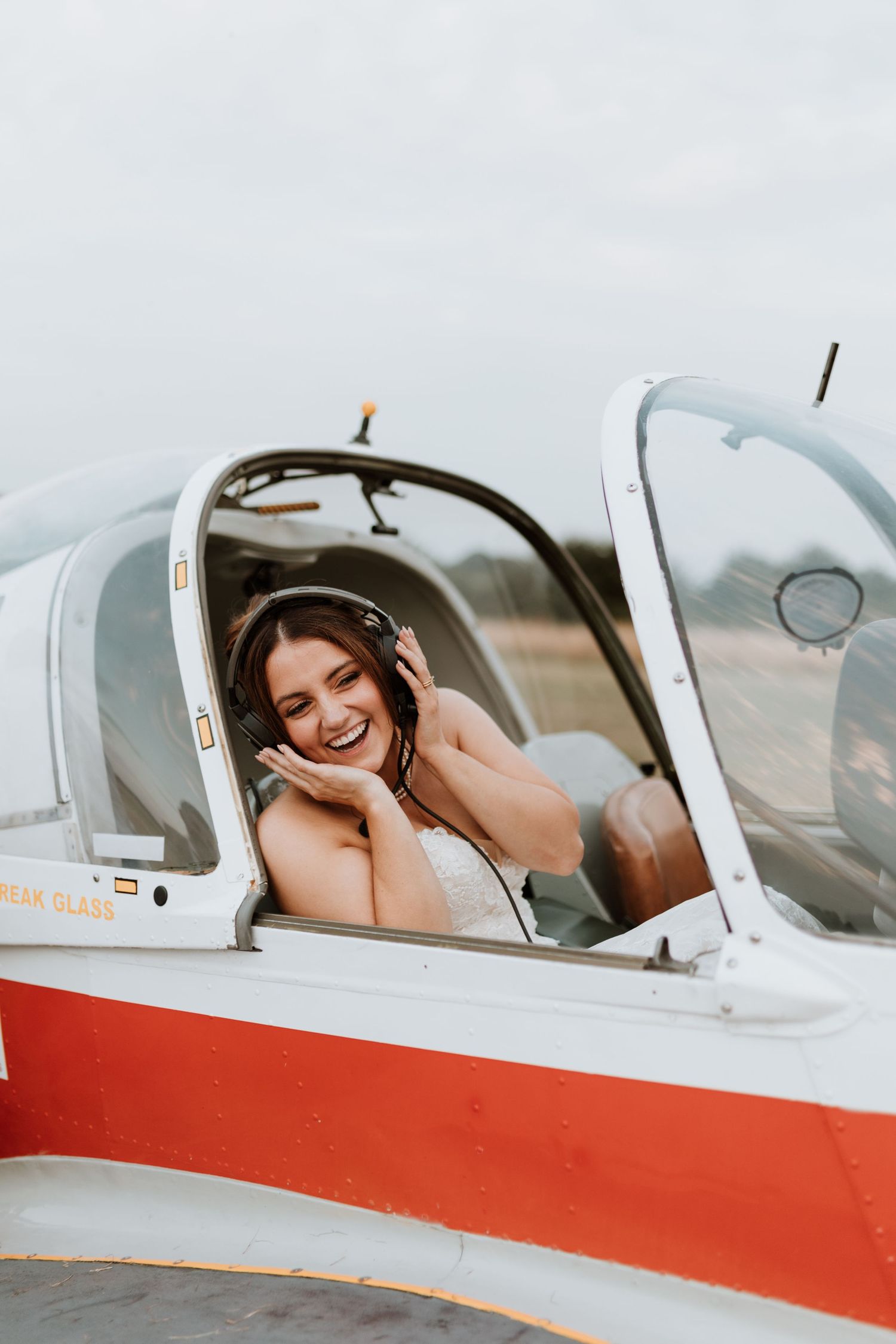 Bride laughs joyfully while sitting in the cockpit of a small white and red airplane.