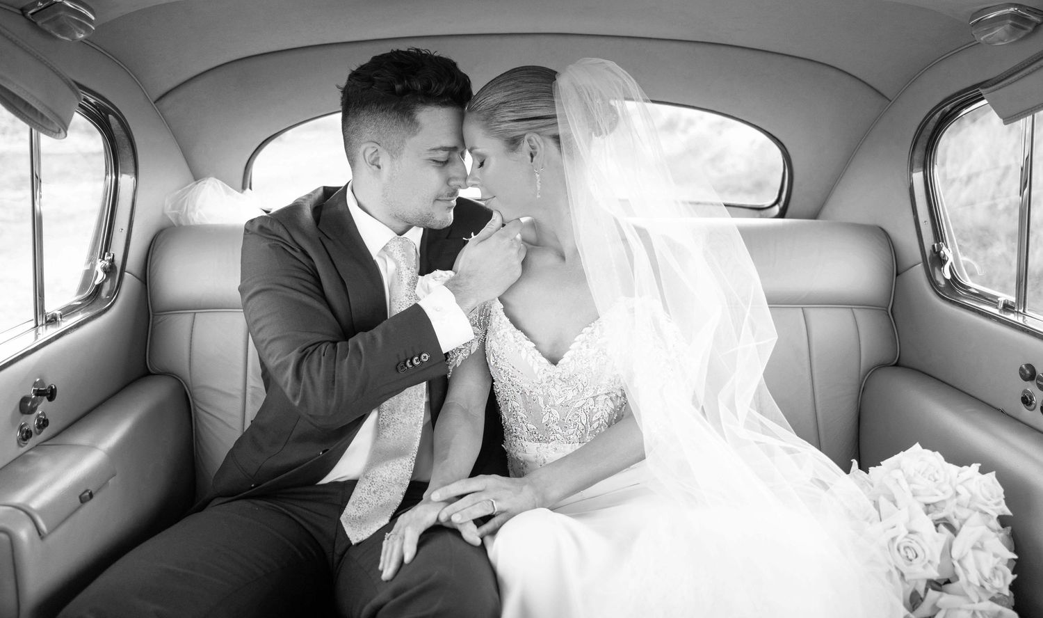 Bride and groom share an intimate moment in the backseat of a vintage car in a black and white wedding photo.
