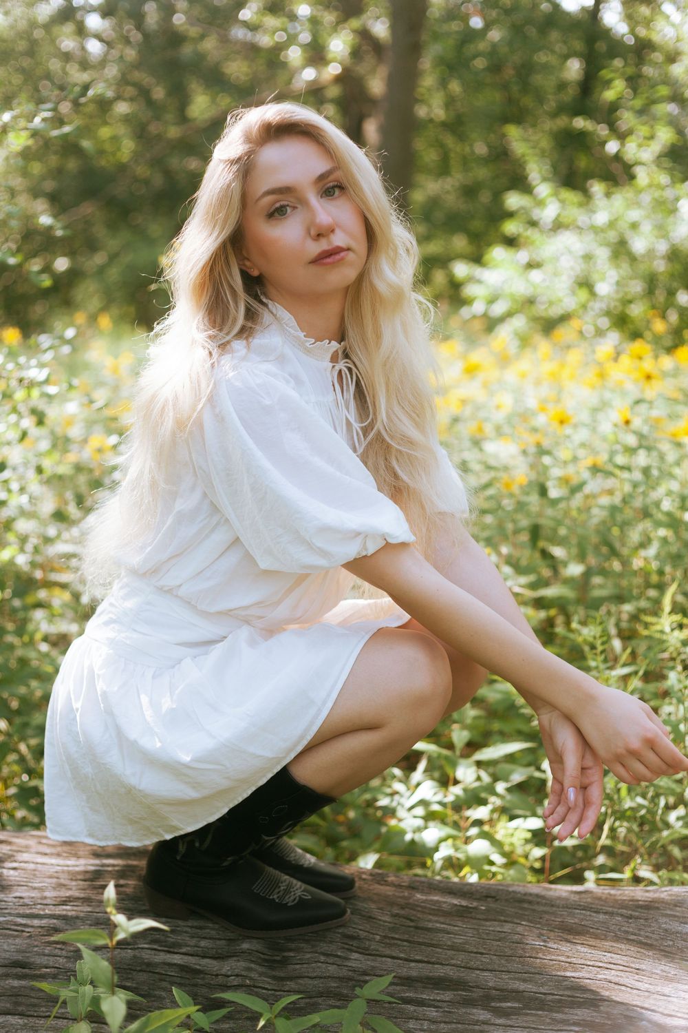 Blonde woman in white dress crouching on log surrounded by yellow wildflowers in a sunlit forest setting.
