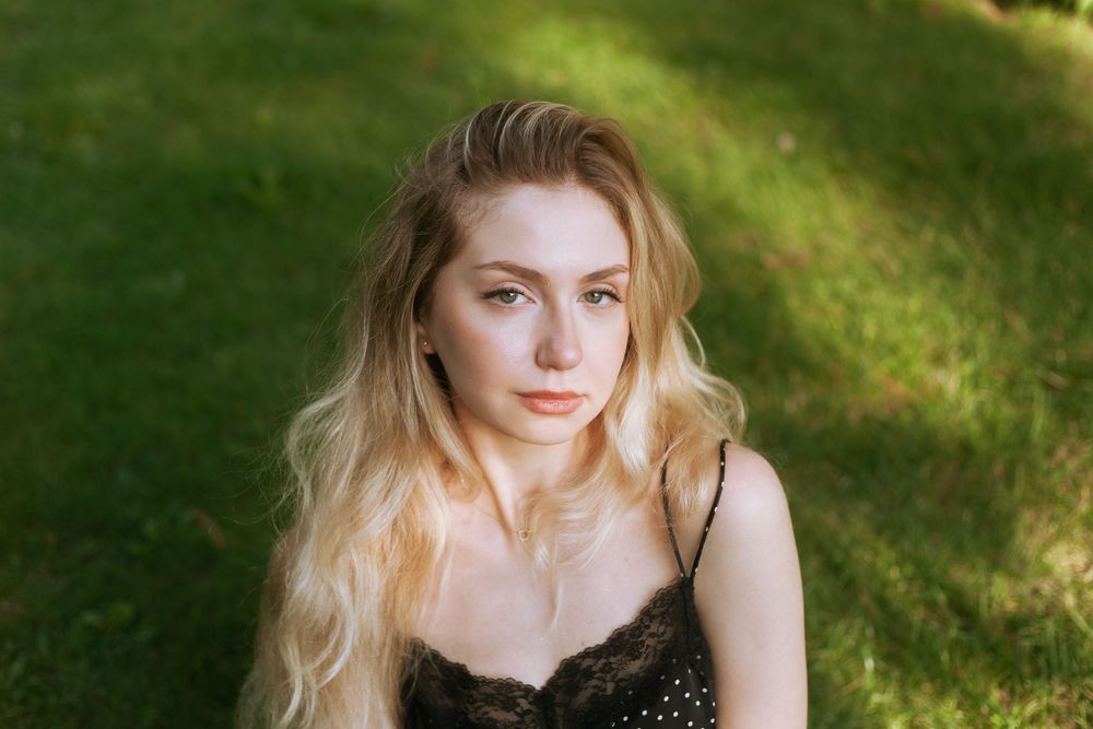 Young blonde woman with wavy hair sitting outdoors on green grass, wearing a black lace top, looking at camera.