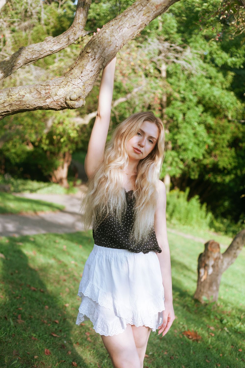 Young blonde woman in black top and white ruffled skirt poses gracefully under a tree branch in a lush green park.