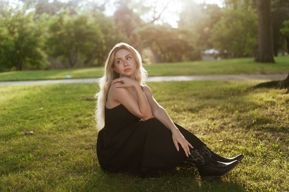 Blonde woman in black dress and boots sitting on green grass in a sunlit park.