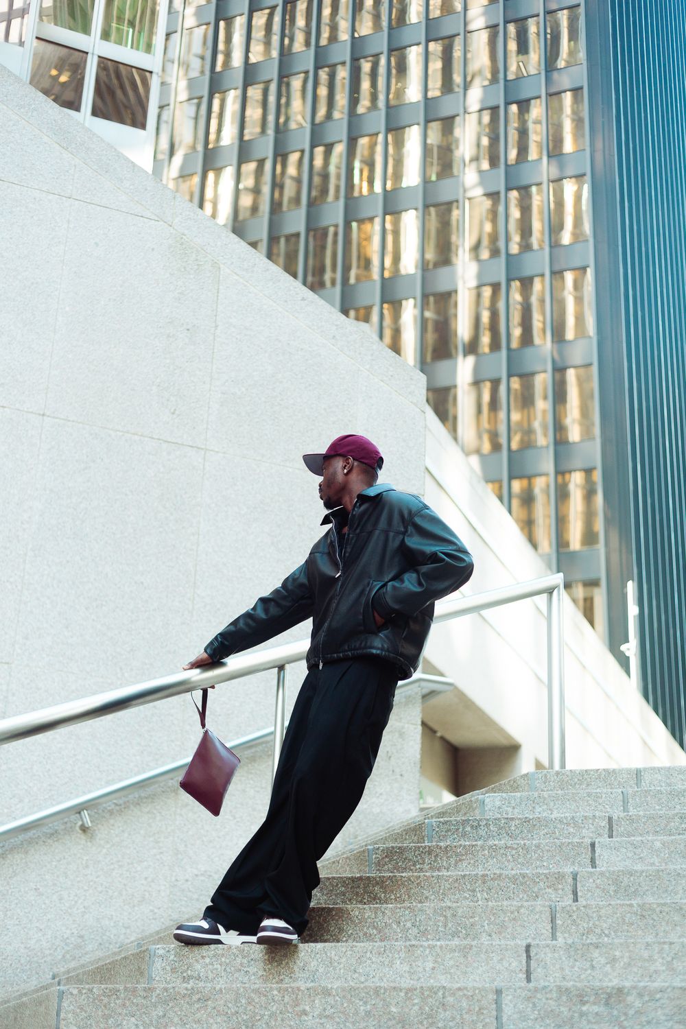 Young Black man in leather jacket and burgundy cap leans on railing of modern urban staircase.