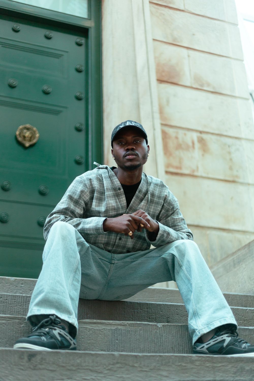 Young Black man in plaid shirt, light jeans and cap sits relaxed on stone steps near green door.