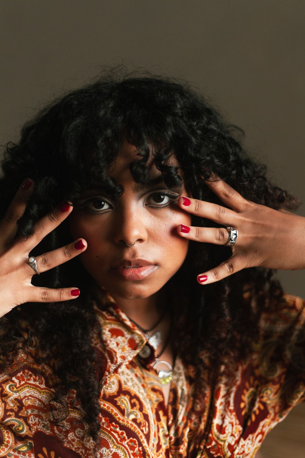 Young Black woman with curly hair poses with red-nailed hands framing her face, wearing a paisley print top.