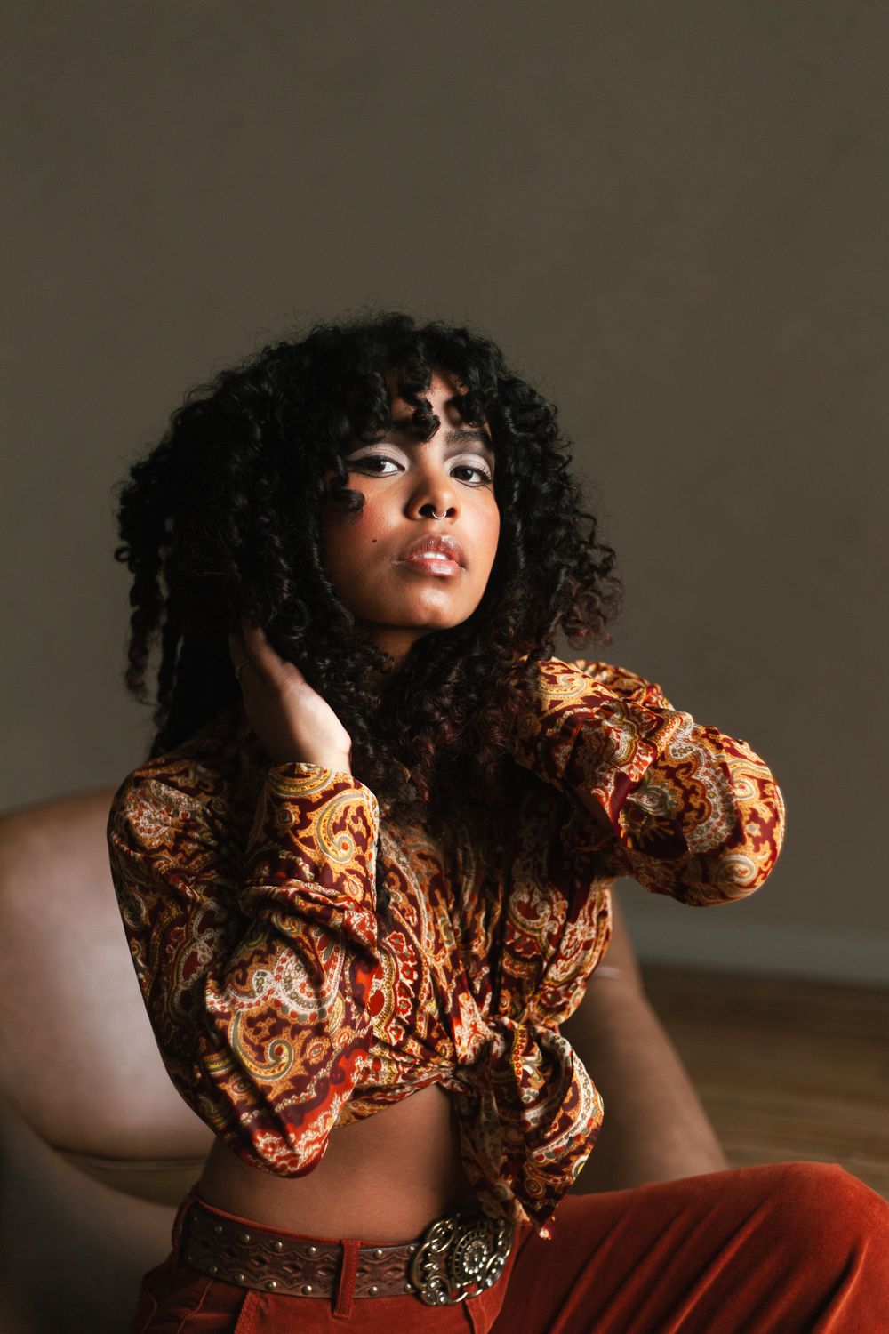 Young Black woman with curly hair sits gracefully in a paisley crop top and rust orange pants in moody lighting.