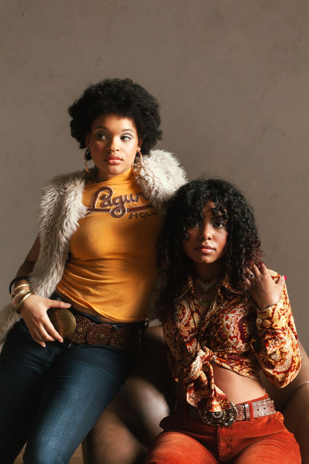 Two young Black women pose in 70s-inspired outfits, one in a yellow tee with fur vest, one in paisley print.