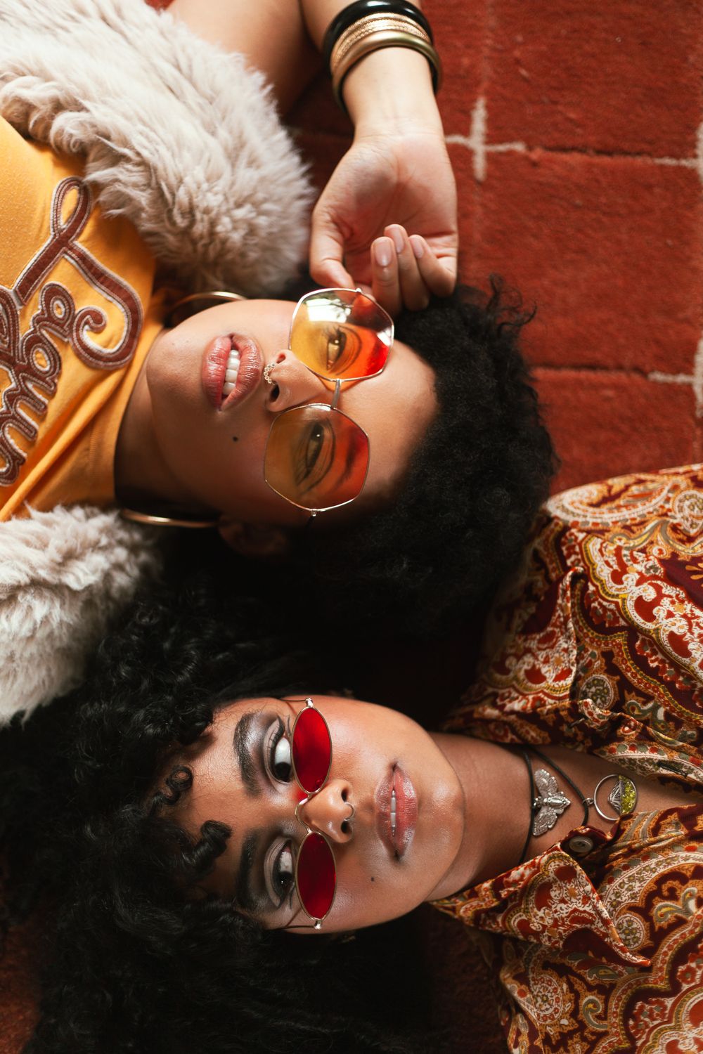 Two young Black women lying down wearing retro tinted sunglasses, styled in 70s-inspired boho fashion.