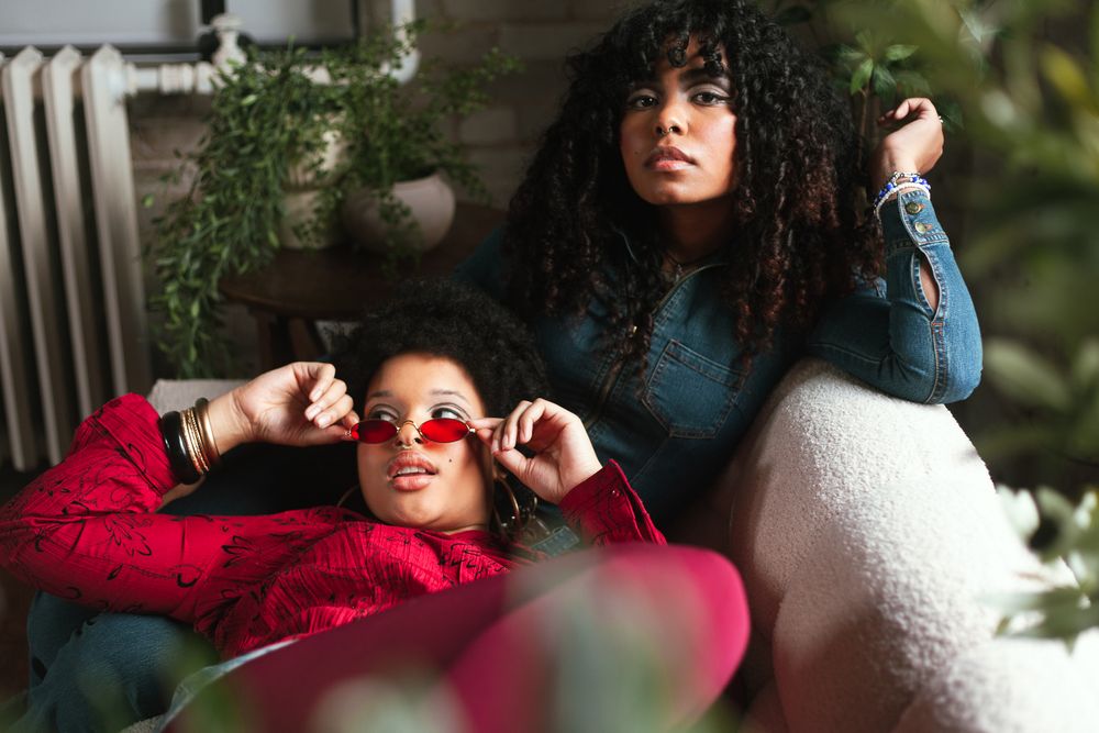 Two stylish young Black women pose together, one in red jacket with sunglasses, other in blue top with curly hair.