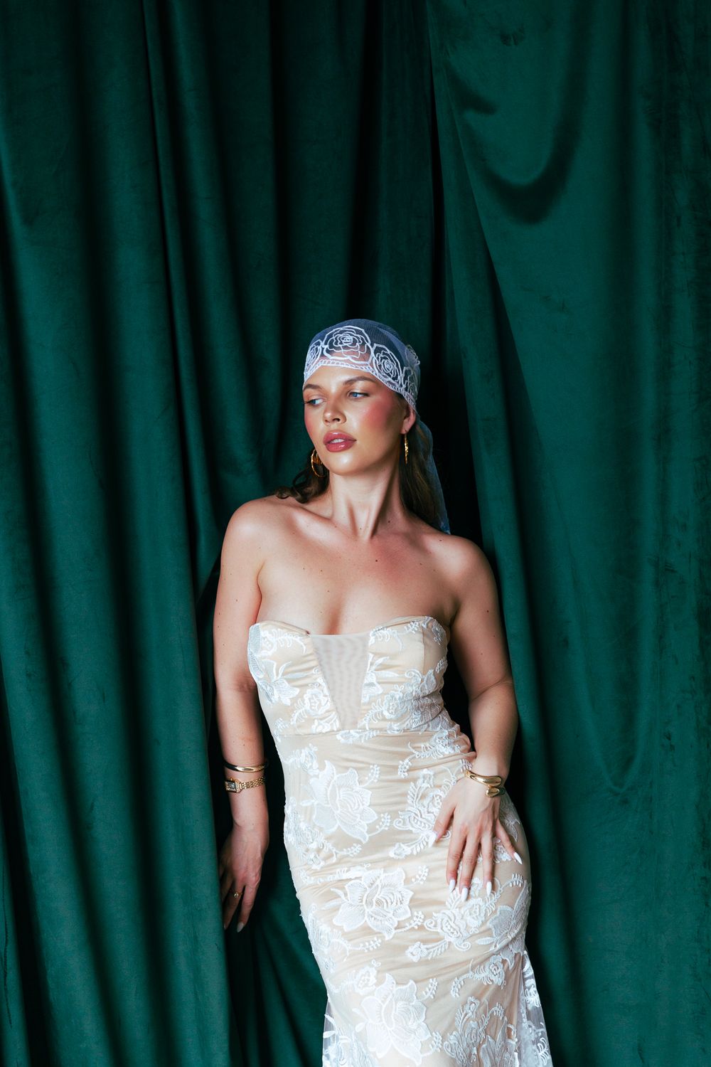 Elegant bride in strapless lace wedding gown with blue lace cap veil against green velvet curtain.