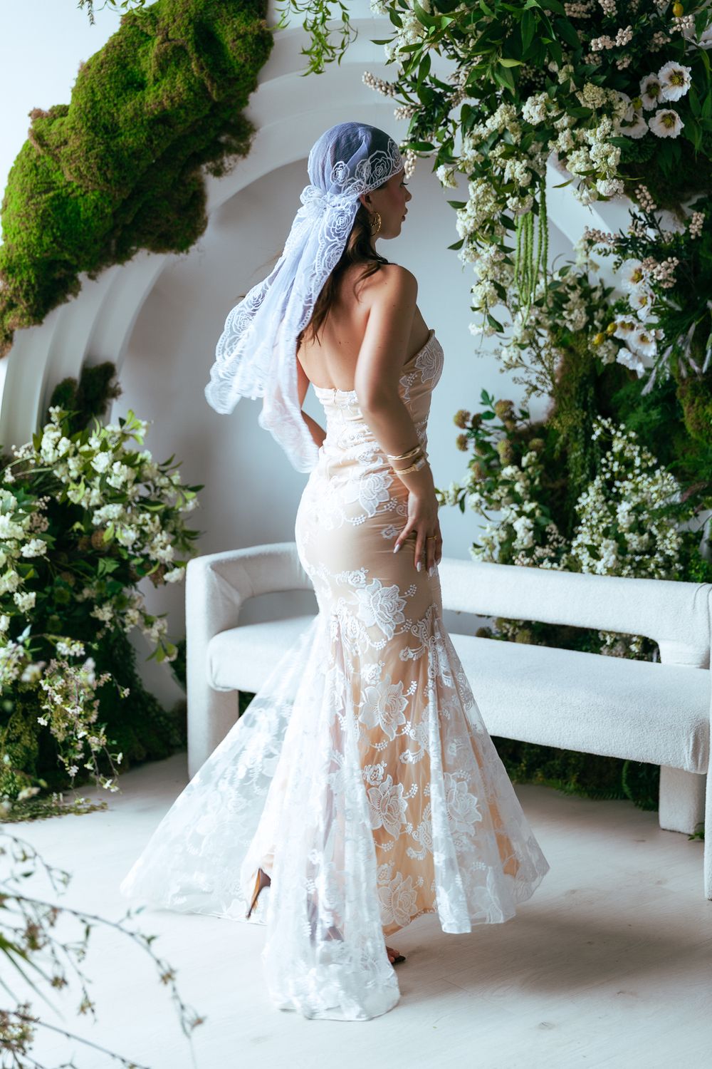 Bride in backless lace mermaid gown with blue veil surrounded by white floral arch decor.