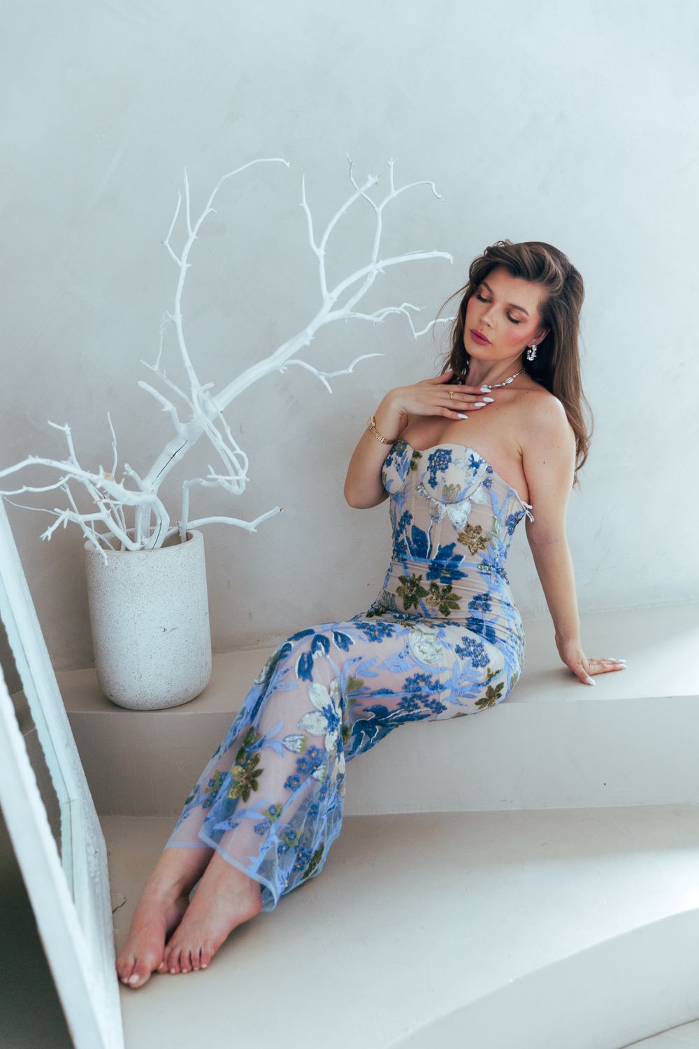 Woman in sheer blue floral strapless gown seated on white steps beside white branch decor.
