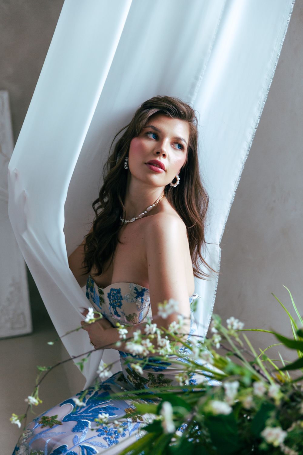 Brunette woman in blue floral strapless gown with pearl necklace among white curtains and flowers.