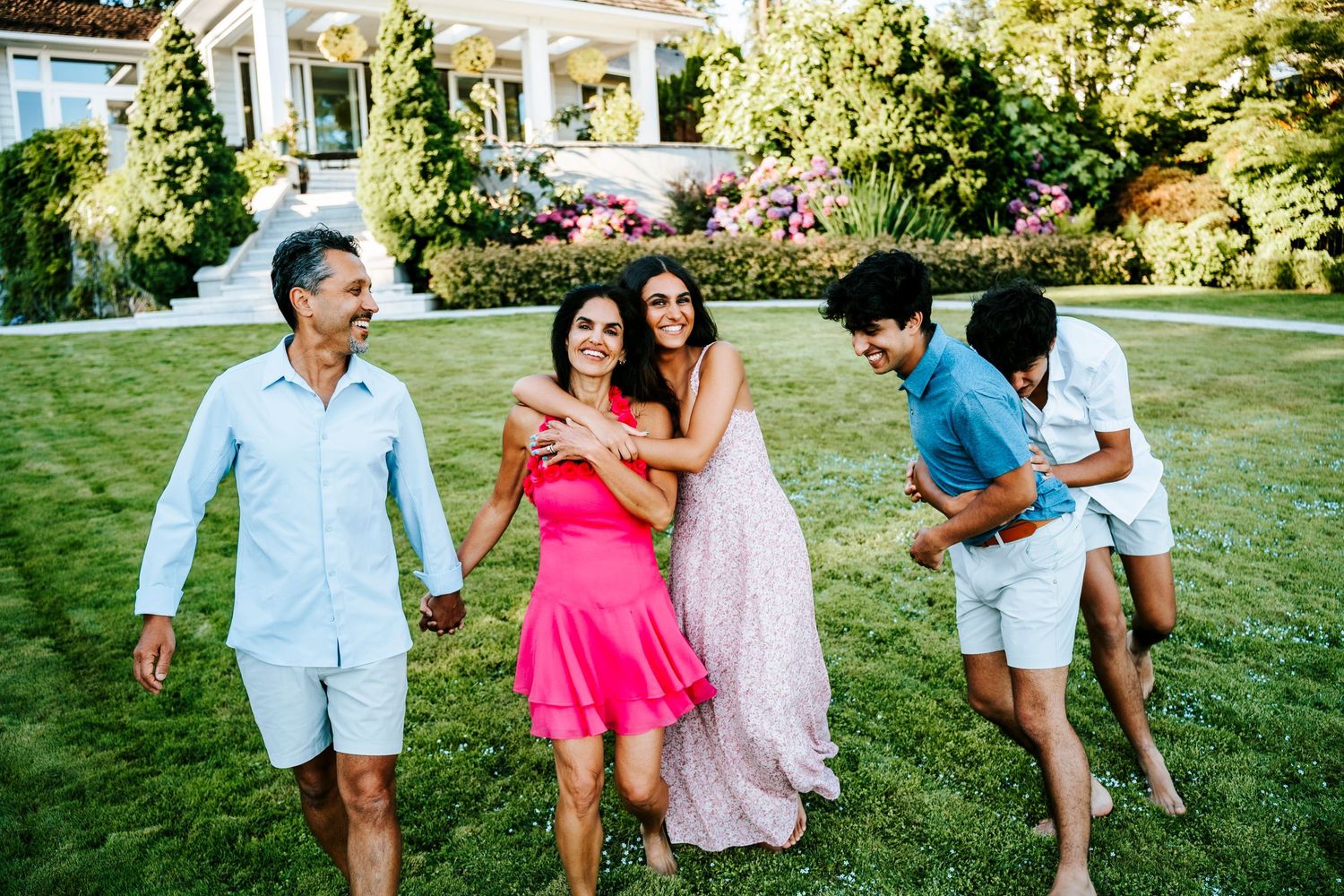 A group of people laughing and dancing together on a lush green lawn outside a white house on a sunny day.