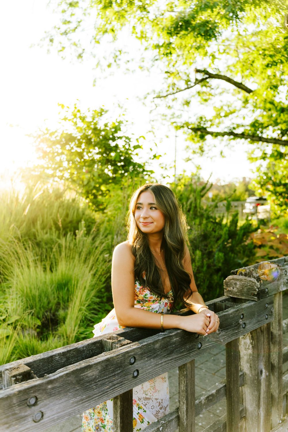 A person in a floral dress leans against a rustic wooden fence with sunlit greenery in the background.