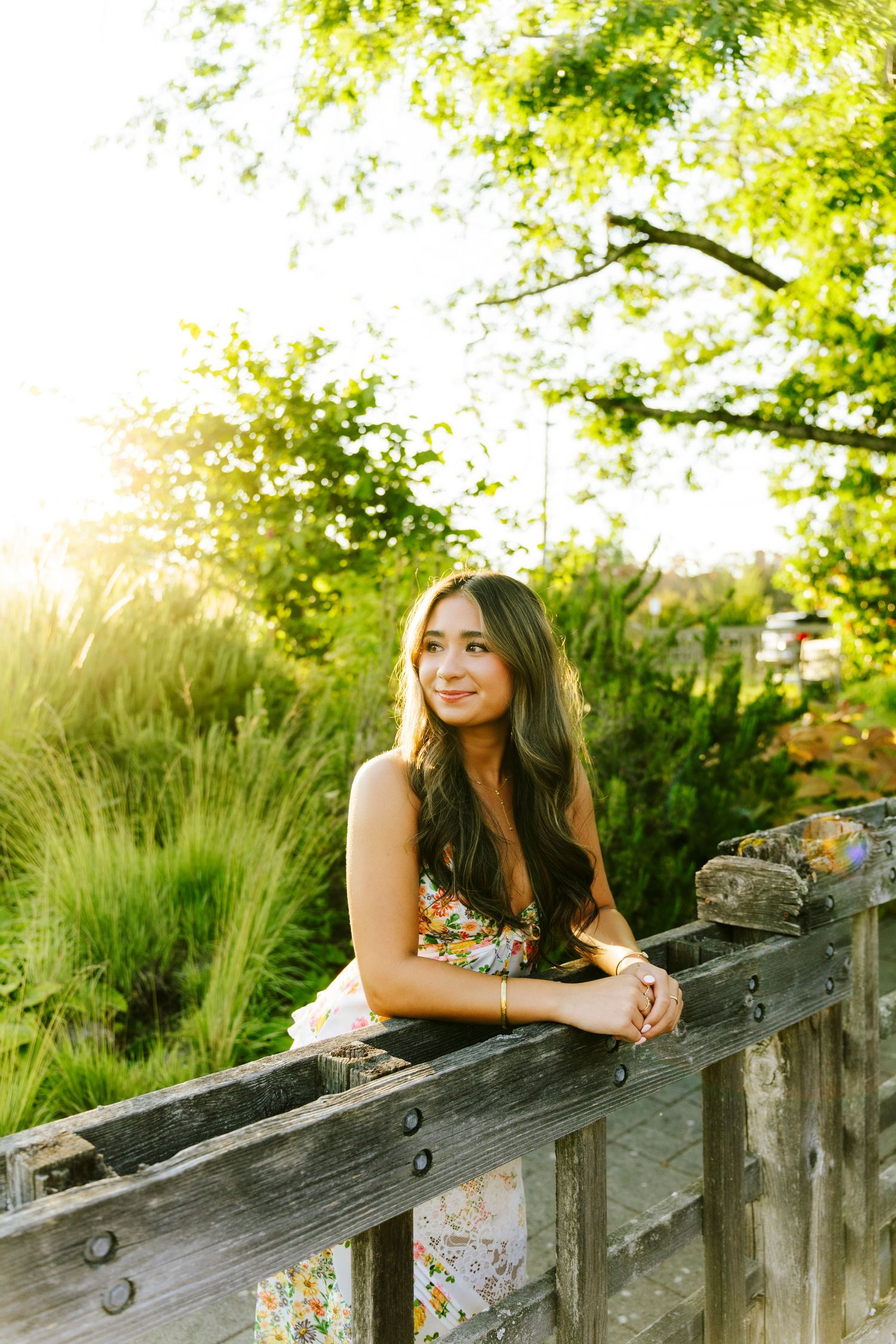 A person in a floral dress leans against a rustic wooden fence with sunlit greenery in the background.