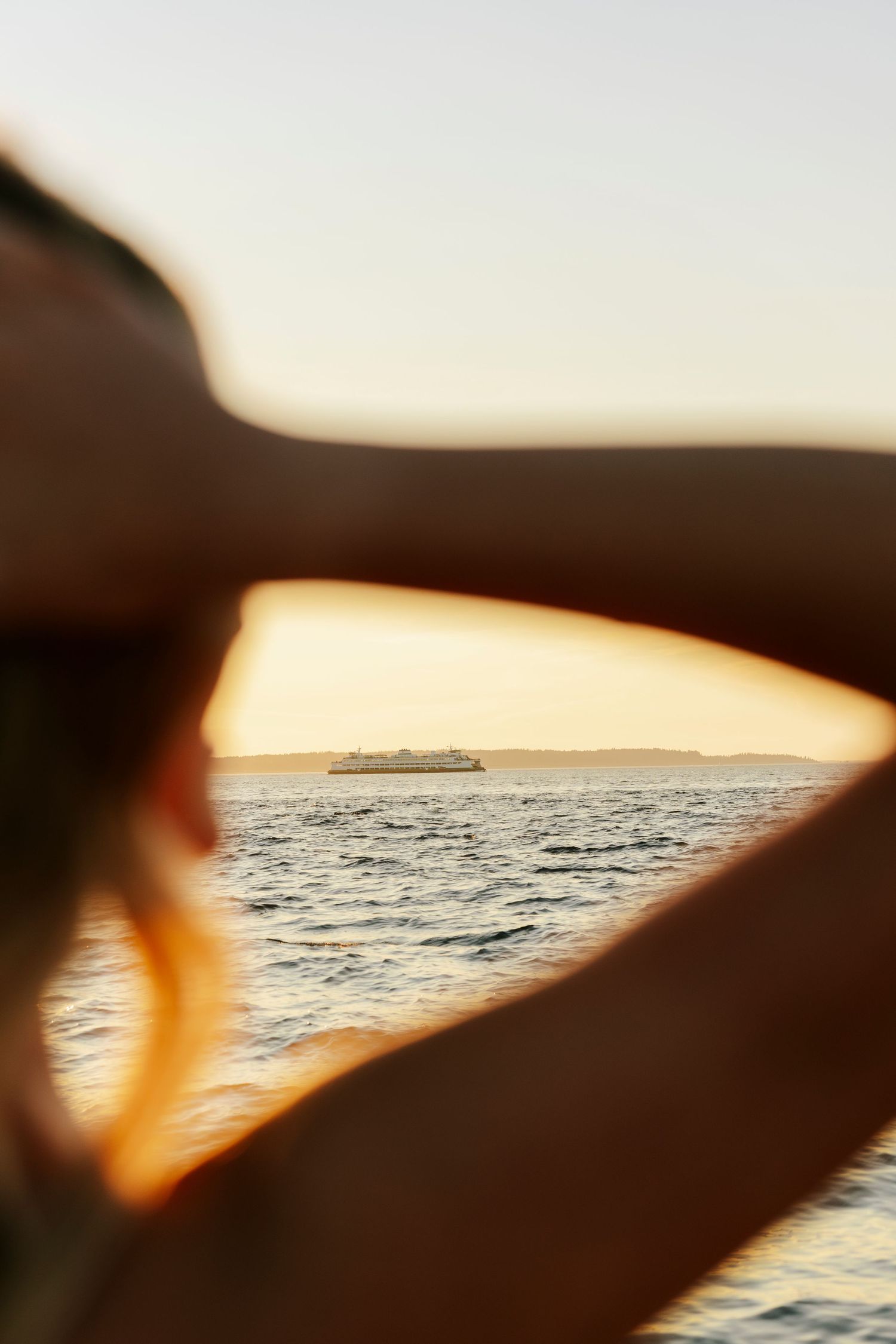 Silhouetted arms frame a distant ship on the ocean horizon during golden hour sunset.