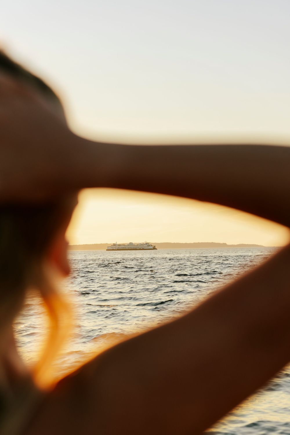 Silhouetted arms frame a distant ship on the ocean horizon during golden hour sunset.