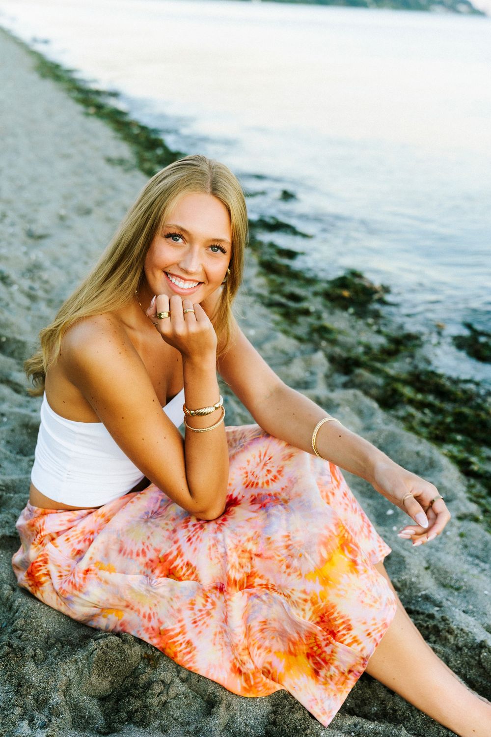 Person in white top and coral tie-dye skirt poses on beach at sunset in series of casual portraits.