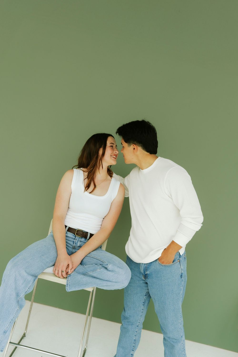 Two people in casual outfits of jeans and white tops pose playfully against a sage green wall.