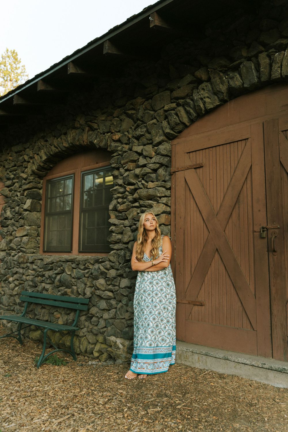 Woman in floral maxi dress leaning against rustic stone building with barn door and bench.