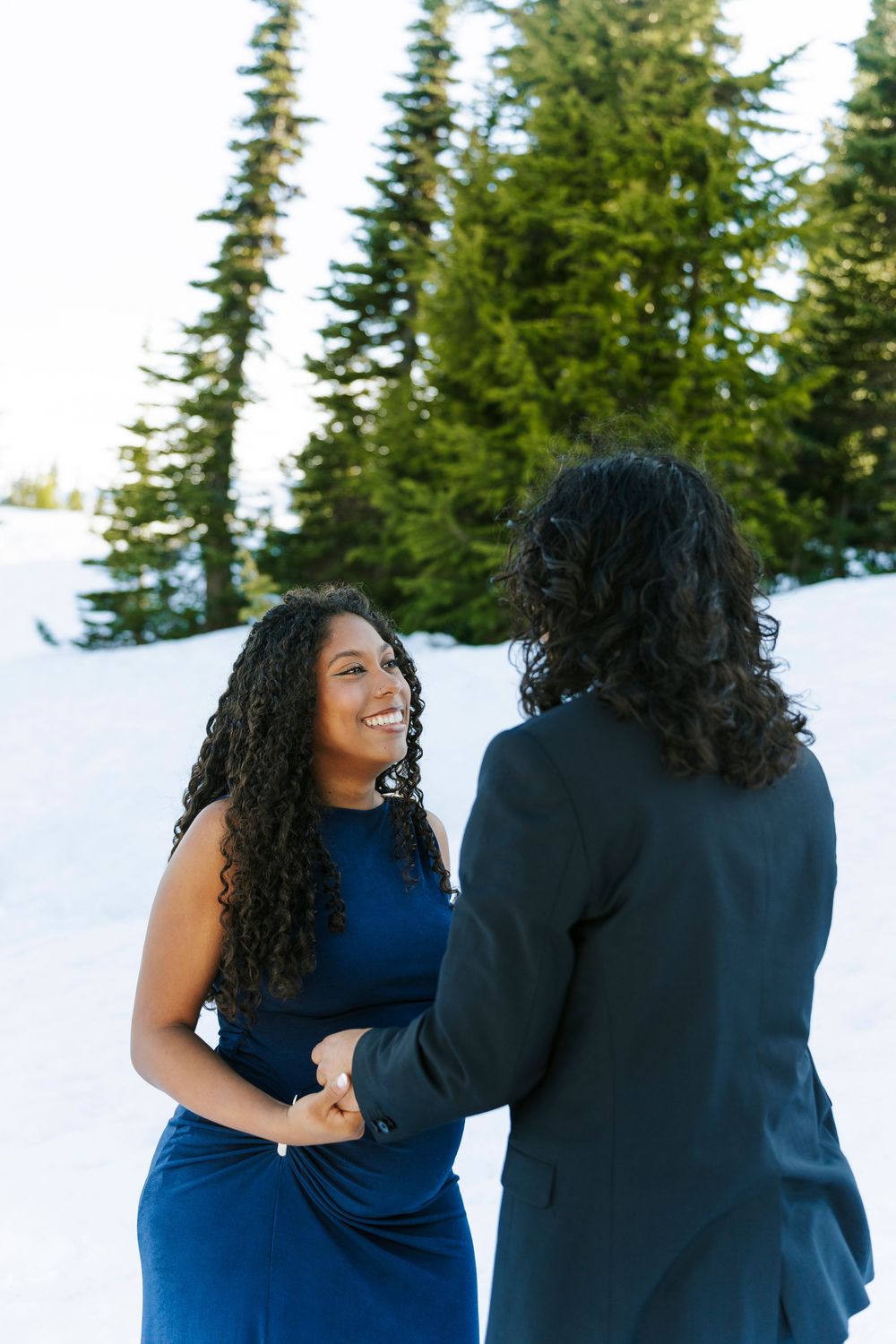 Two women holding hands in the snow, one in a blue dress smiling, with evergreen trees behind them.