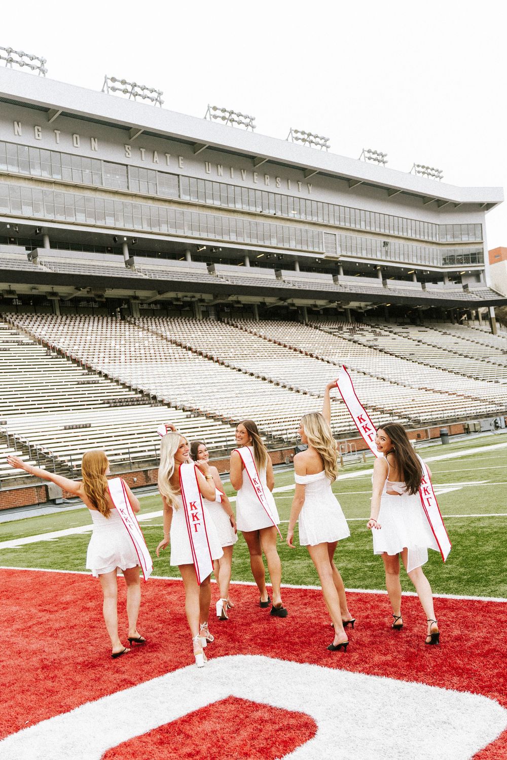 Five women in white dresses and red sashes pose on a college football field at what appears to be Kent State University.