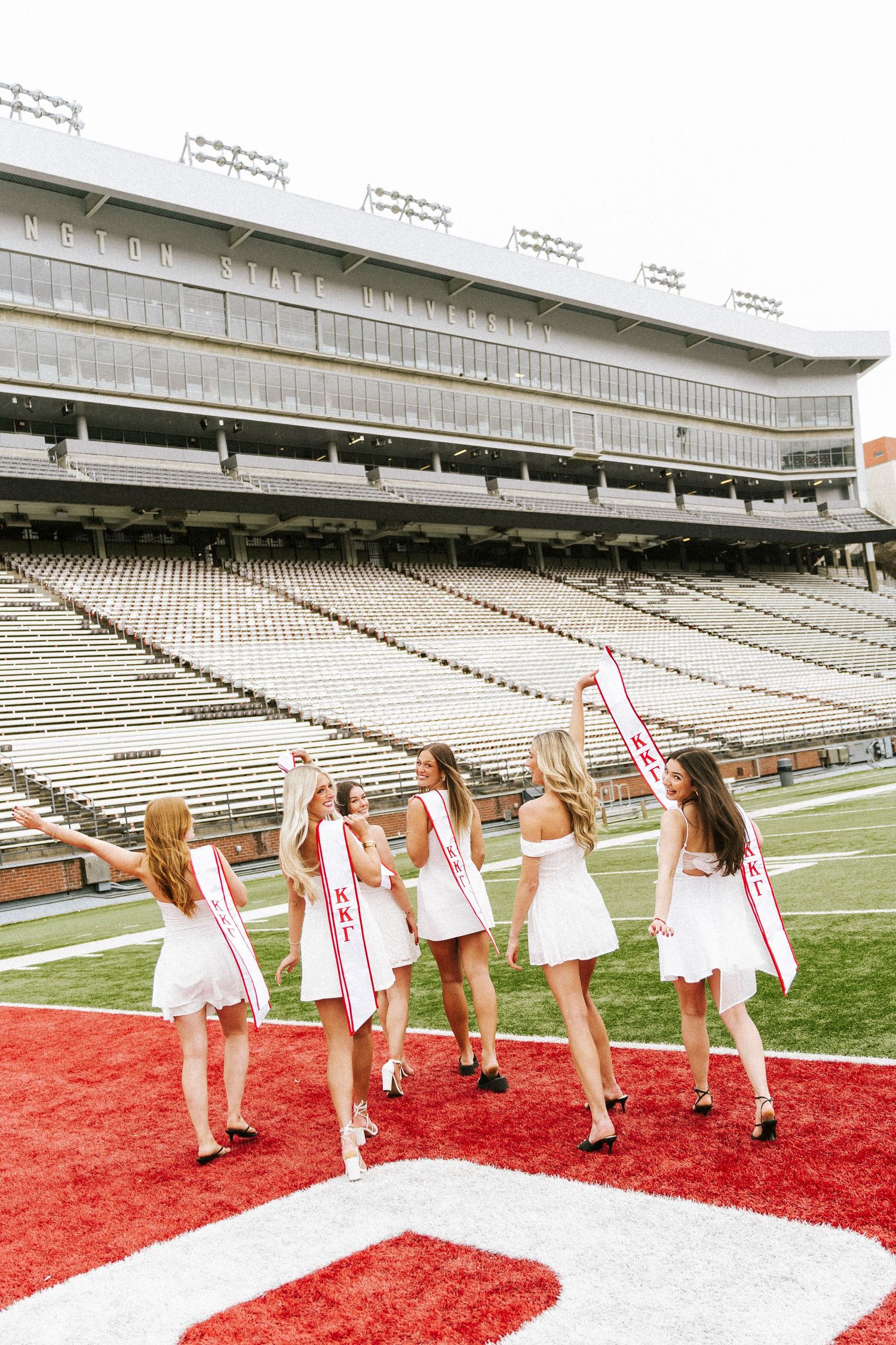 Five women in white dresses and red sashes pose on a college football field at what appears to be Kent State University.
