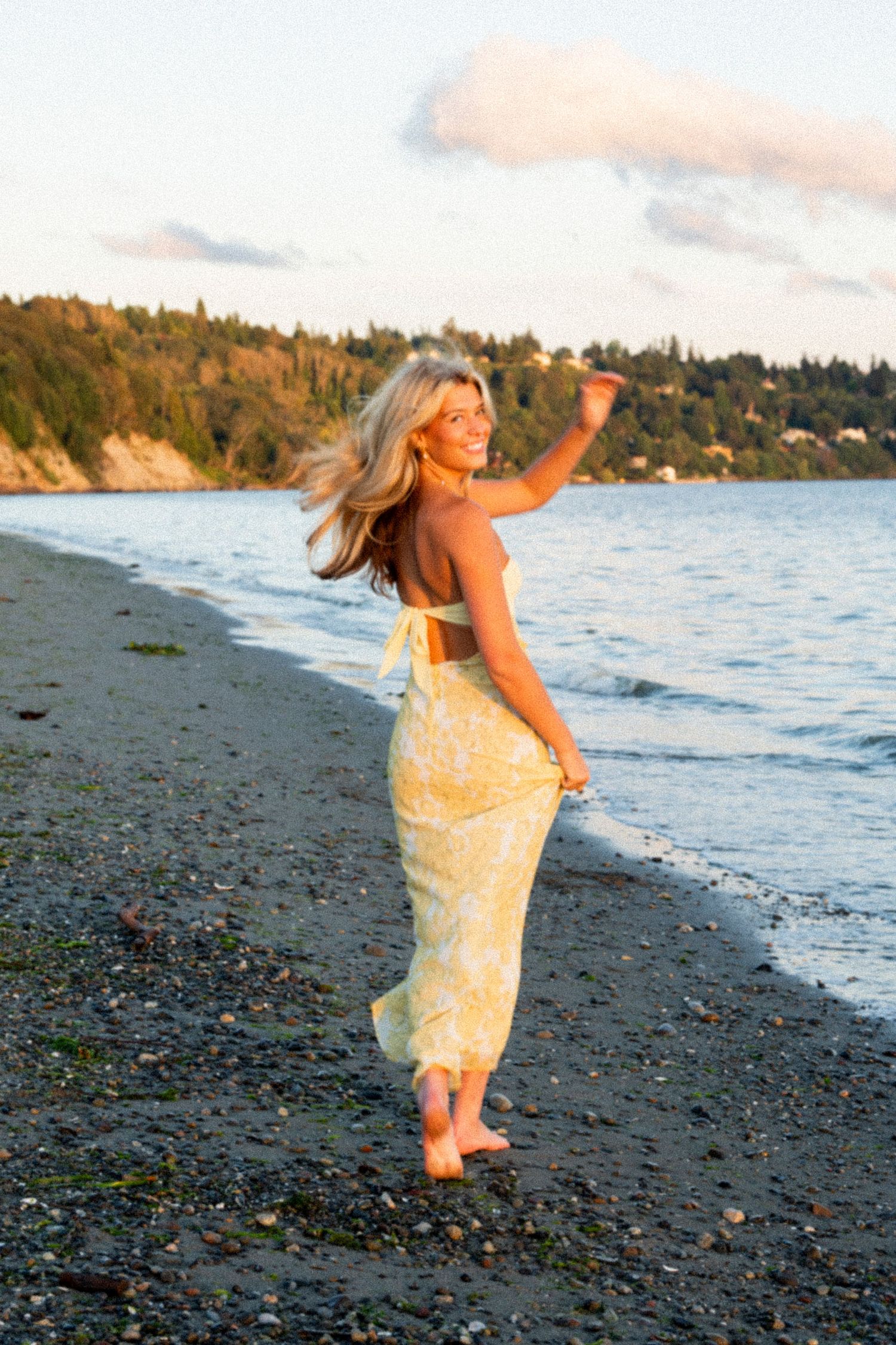 A woman in a yellow sundress dances joyfully on a rocky beach at sunset with water and trees in the background.