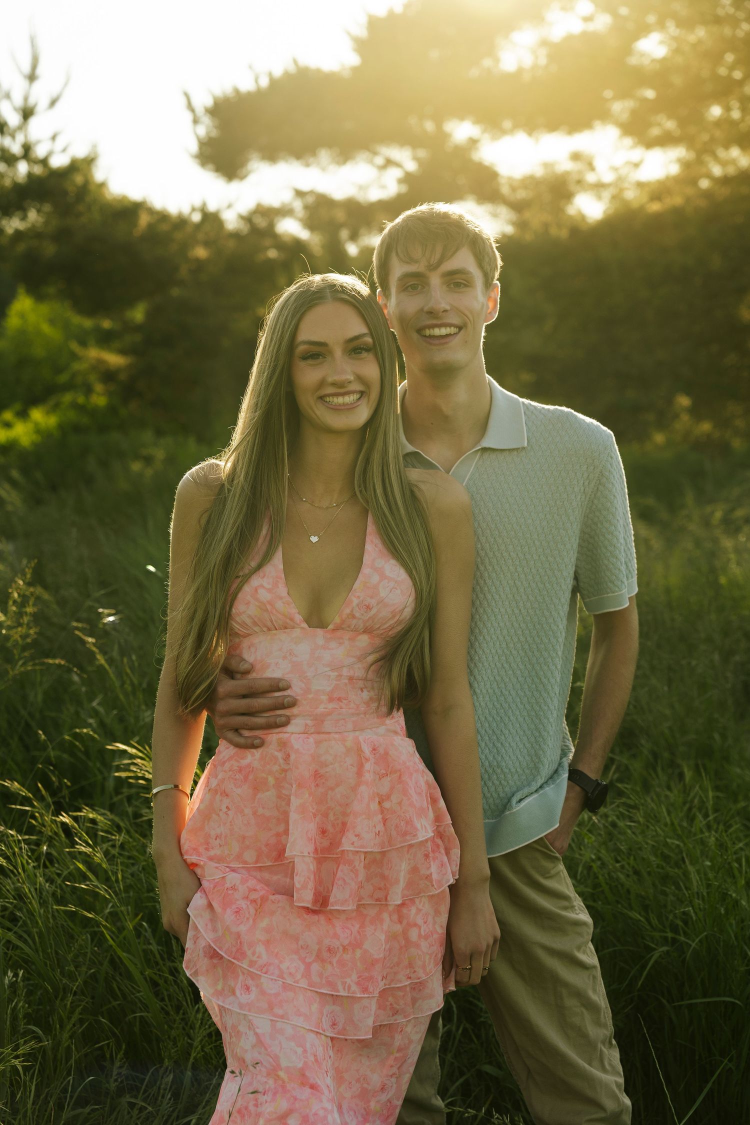A young couple smiles outdoors at golden hour, woman in pink floral dress, man in light polo shirt, lush greenery background.