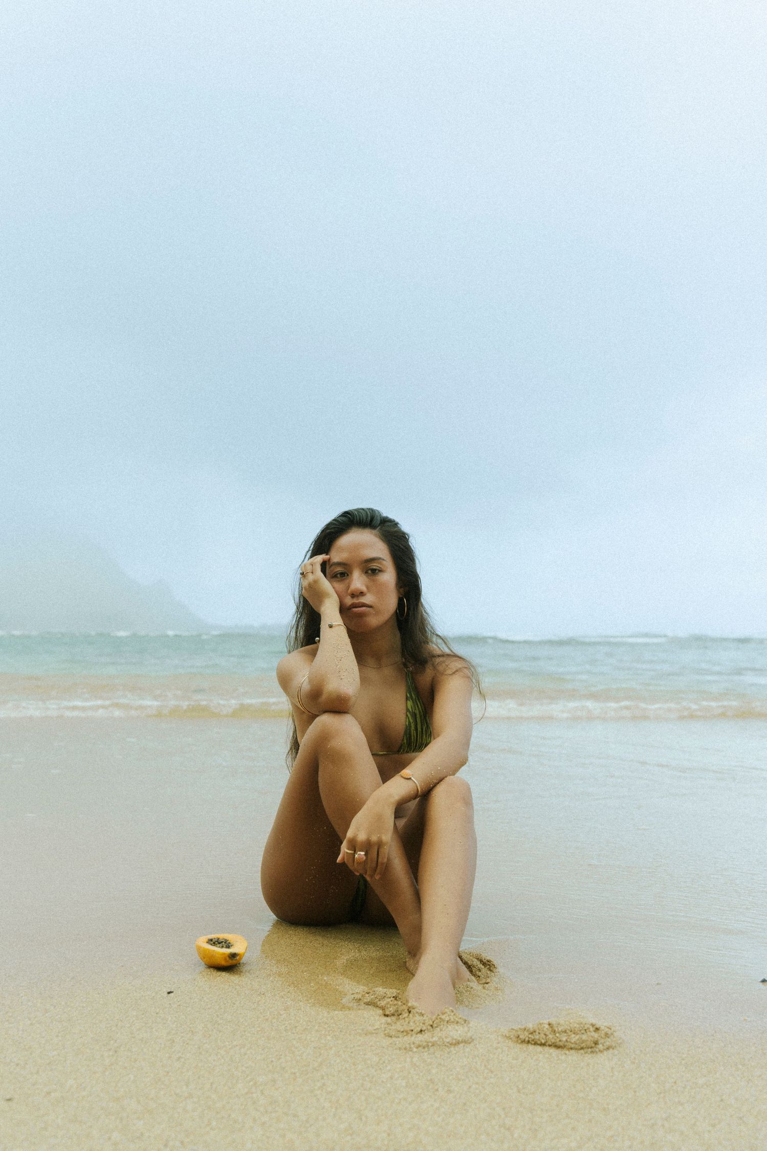 Woman in bikini sitting on sandy beach shore with ocean waves and misty mountains in background.
