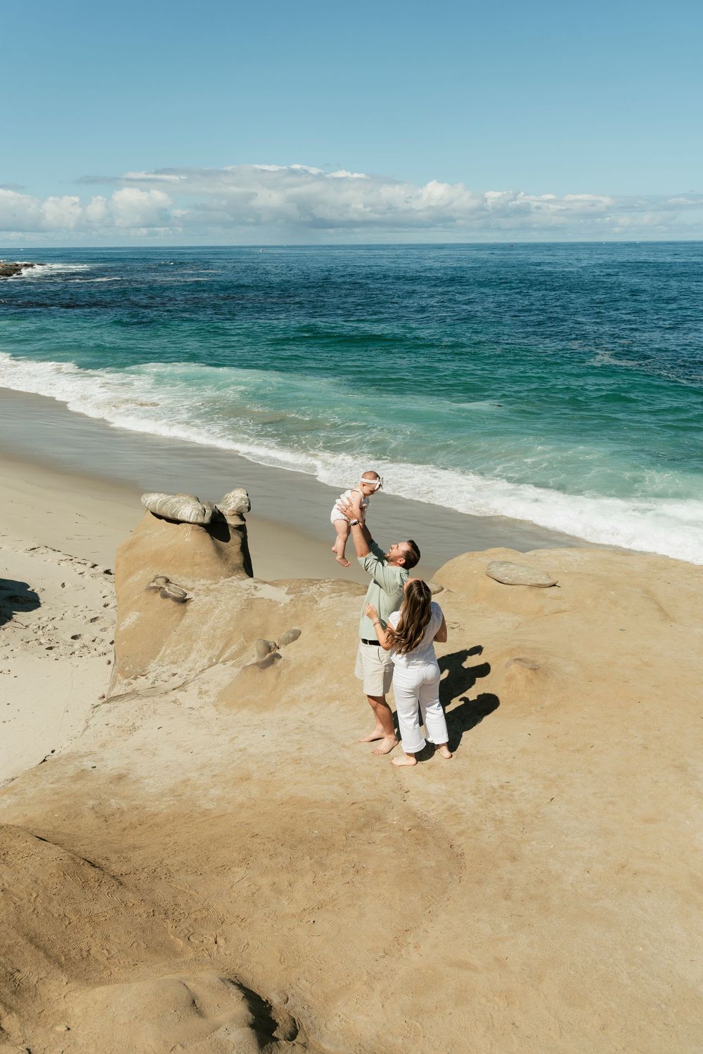 Family enjoying a sunny day at the beach, with a parent lifting a baby in the air near the ocean waves.