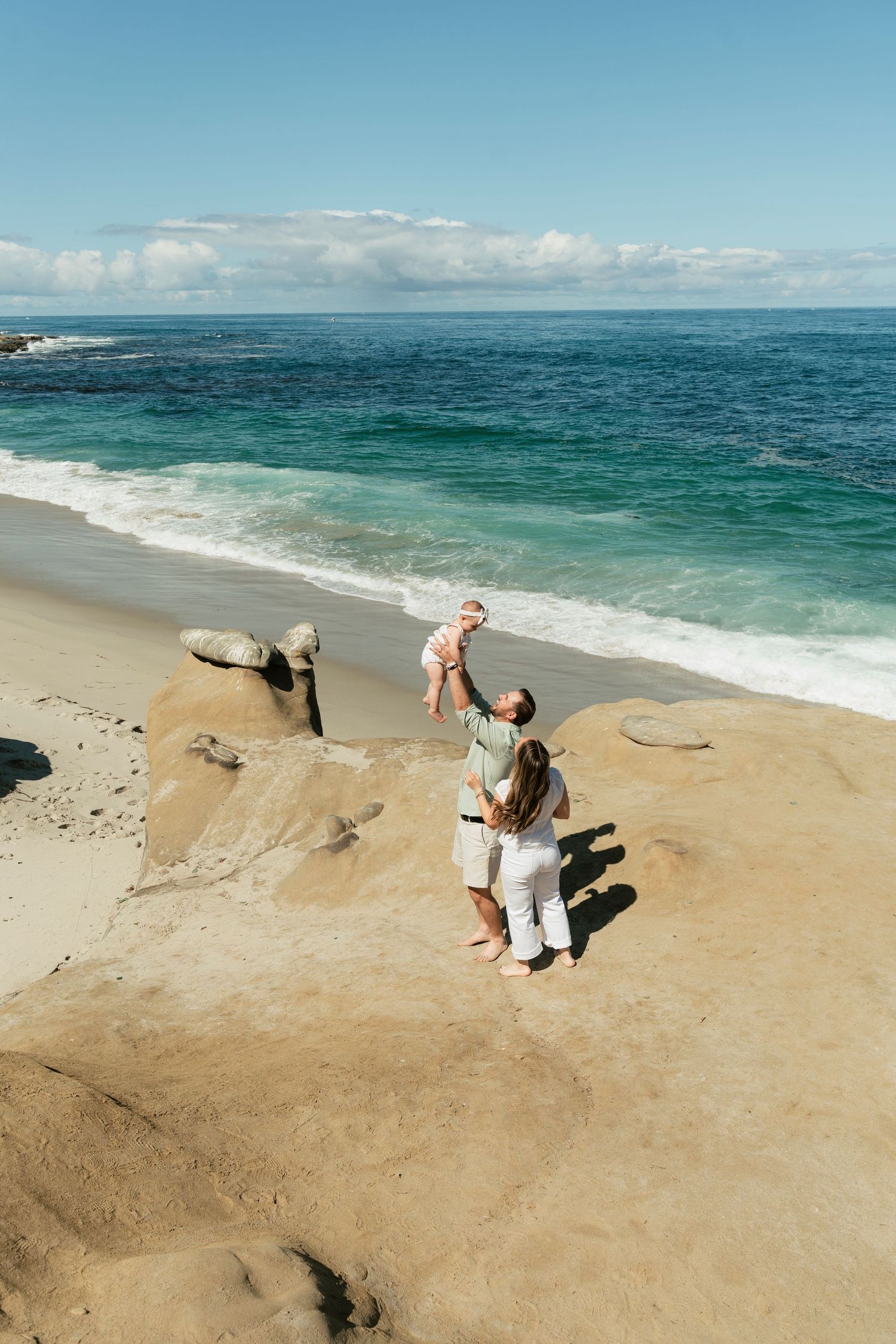Family enjoying a sunny day at the beach, with a parent lifting a baby in the air near the ocean waves.