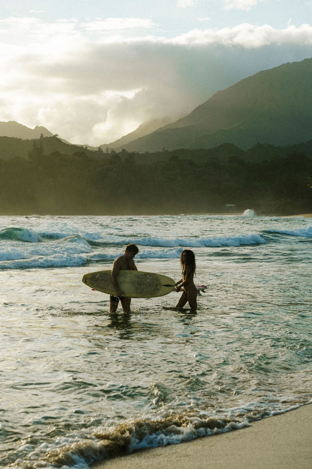 Two surfers wade through golden ocean waves at sunset with misty mountains in the background on a tropical beach.