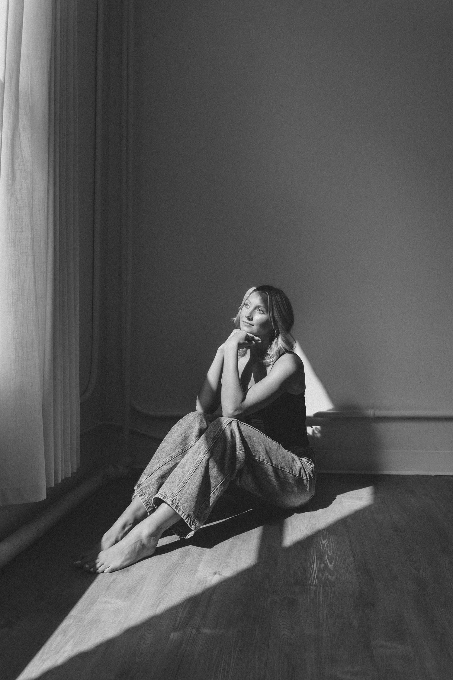Black and white photo of a woman sitting barefoot on a hardwood floor in sunlight, gazing upward thoughtfully.