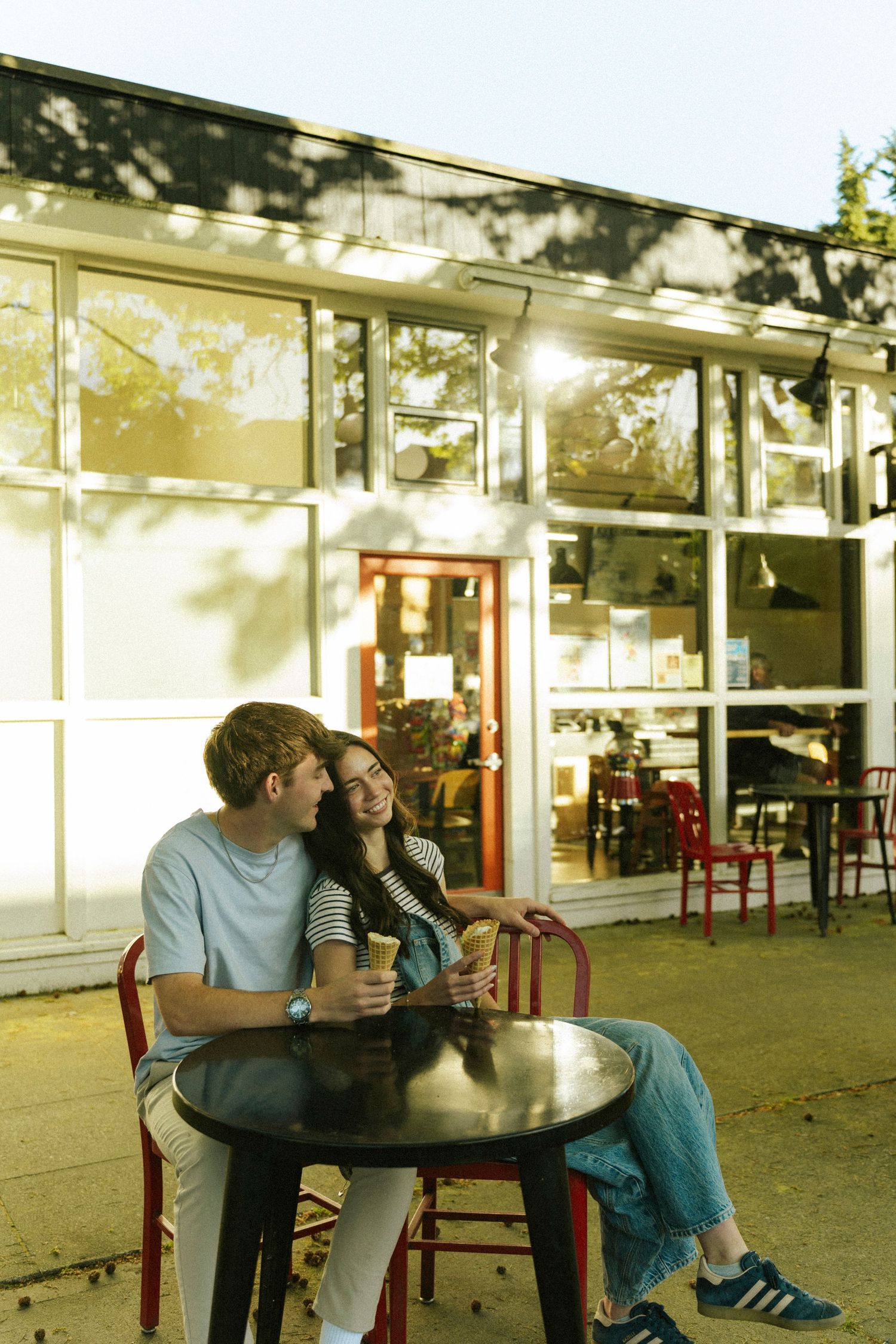 A couple shares ice cream at an outdoor cafe table, smiling at each other in warm golden sunlight.
