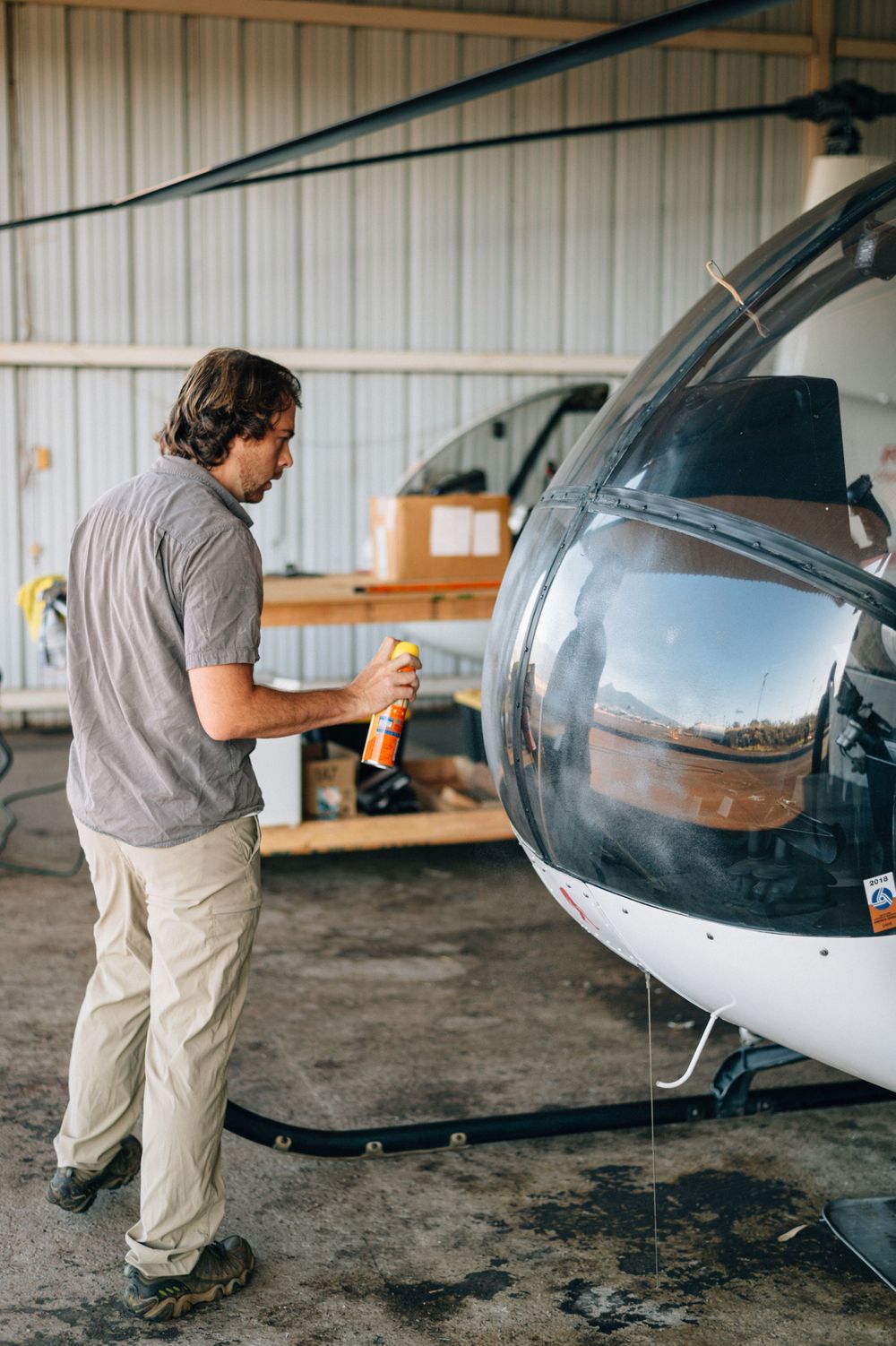 Maintenance being performed on a small silver helicopter inside a hangar.