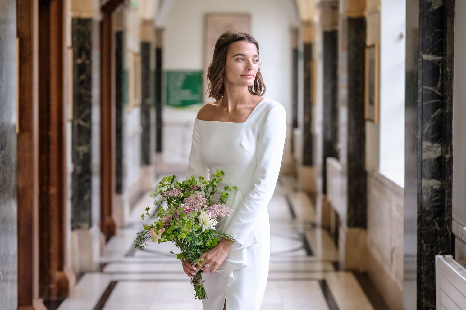 A bride in a long-sleeved white wedding gown walks through an elegant hallway holding a green and white bouquet at Islington Town Hall