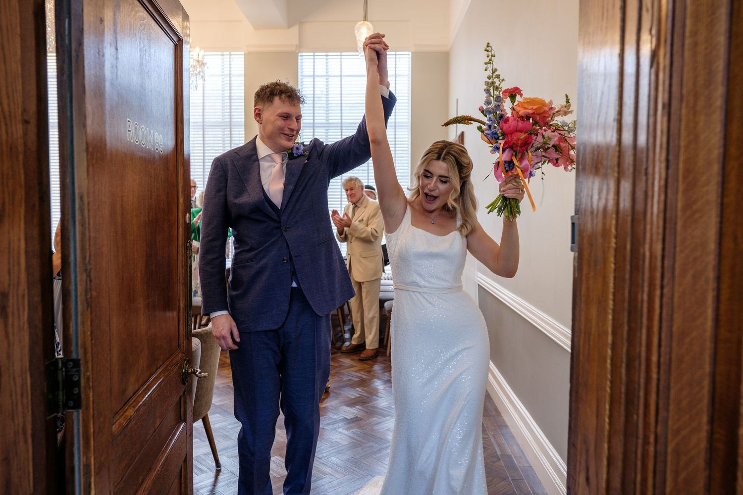 A joyful couple celebrates their wedding day as they walk through a hallway with raised arms and flowers at Islington Town Hall