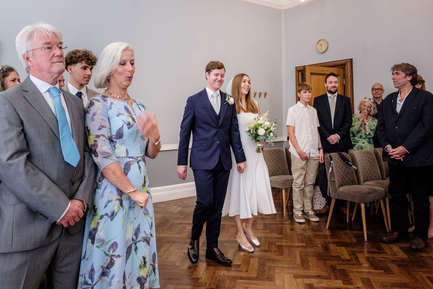 A group of wedding guests gathered in a Room 99 at Islington Town Hall with wooden floors and white walls during a ceremony.