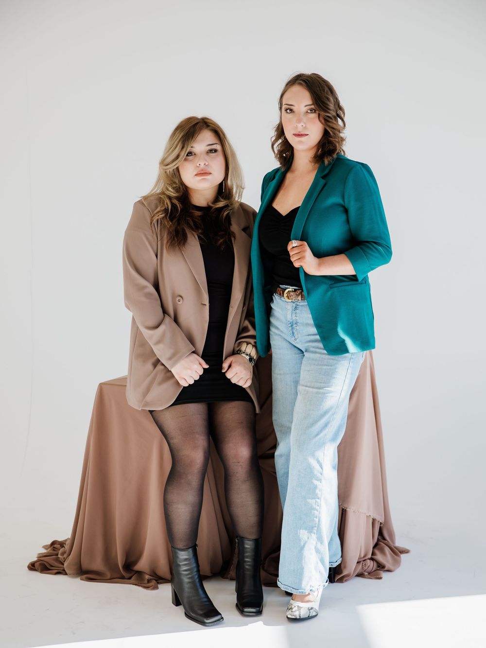 Two people in contrasting outfits of teal blazer and brown coat pose together against white background.