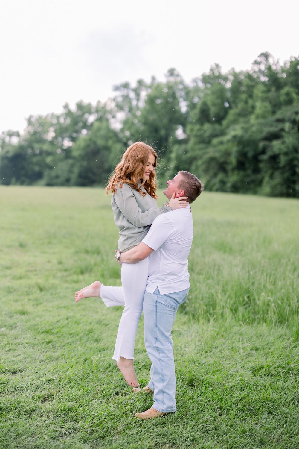 Playful couple dancing and embracing in a lush green field surrounded by trees.